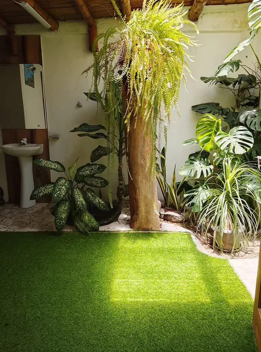 Patio interior con césped artificial, plantas tropicales y un tronco de árbol, con el área del lavabo a la izquierda