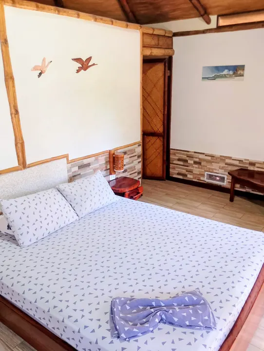 Cama grande con sábanas blancas estampadas con hojas, toalla lila en forma de lazo, puerta de madera con marco de bambú y techo de madera