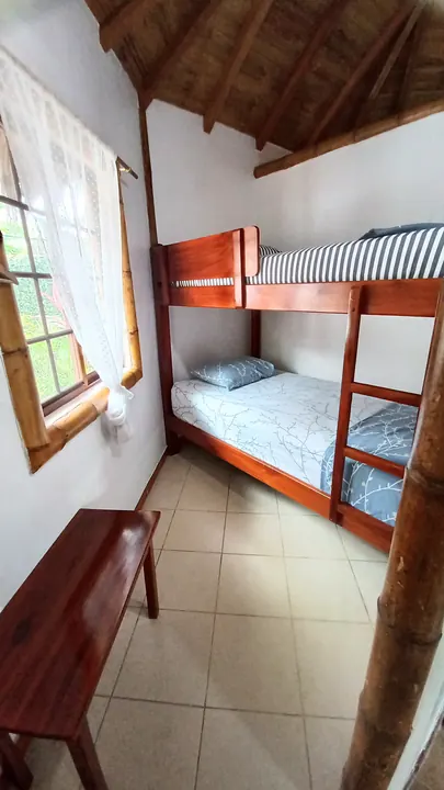 Cama litera de madera en la cabaña familiar, con ropa de cama a rayas y gris, ventana con cortina de encaje y techo de paja