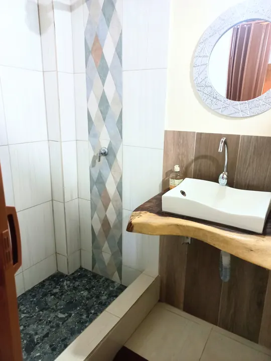 Baño de la cabaña familiar 6 con ducha de azulejos en rombos, piso de piedras oscuras y lavabo blanco sobre madera natural