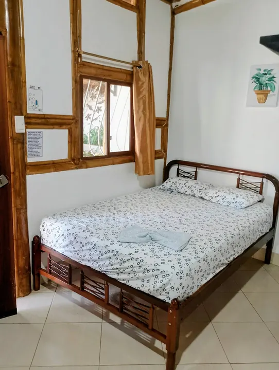 Cama de dos plazas con sábanas blancas floreadas, ventana de bambú con cortina y piso de cerámica