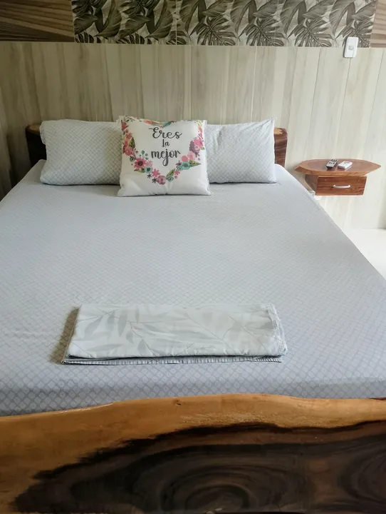 Cama amplia con sábanas grises, cojín floral con la frase «Eres la mejor», pie de cama de madera natural y mesita de noche flotante