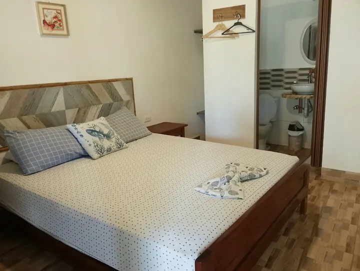 Cama queen con cabecera de madera en espiga, almohadas azules y floreadas, y la puerta del baño abierta al fondo