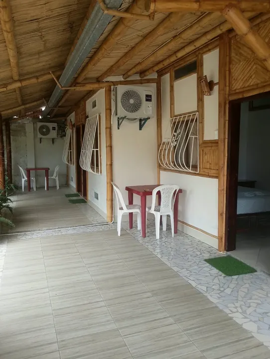 Corredor techado de bambú frente a las habitaciones, con mesas rojas, sillas blancas y equipos de aire acondicionado